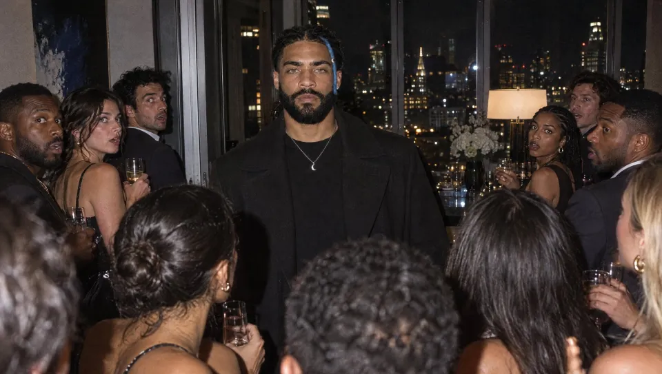 Standing still and unsmiling in the middle of a crowded penthouse party, guests with drinks turning to look at him.
