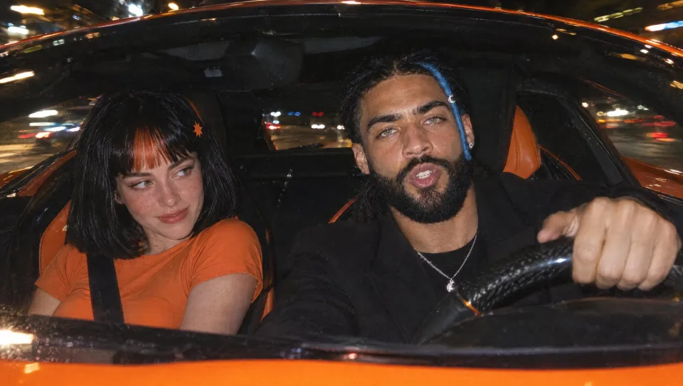 Seen through the windshield, he drives the orange car at night while Claudia in her orange tee watches him from the passenger seat.