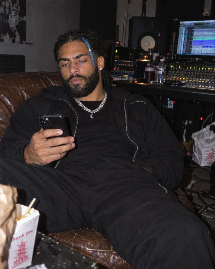 Slouched on a worn leather couch in a recording studio, reading his phone, a takeout carton in the foreground and the mixing desk behind.