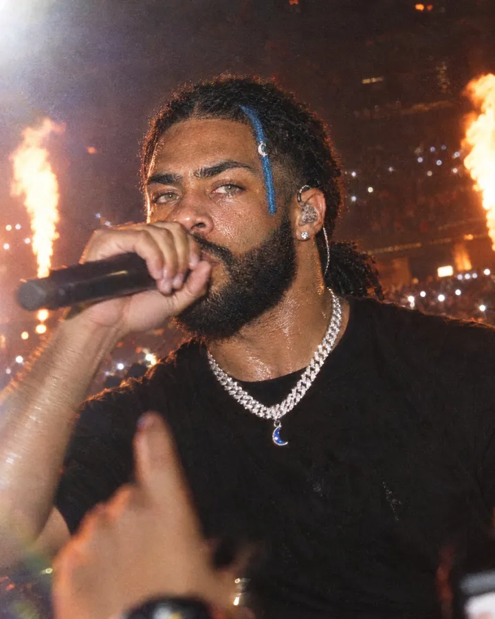 On stage, rapping into a handheld mic with an in-ear monitor, flames shooting up and crowd lights behind.